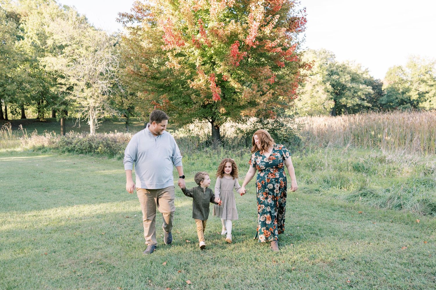 Family of four walking hand-in-hand through a green meadow with colorful autumn trees in the background.