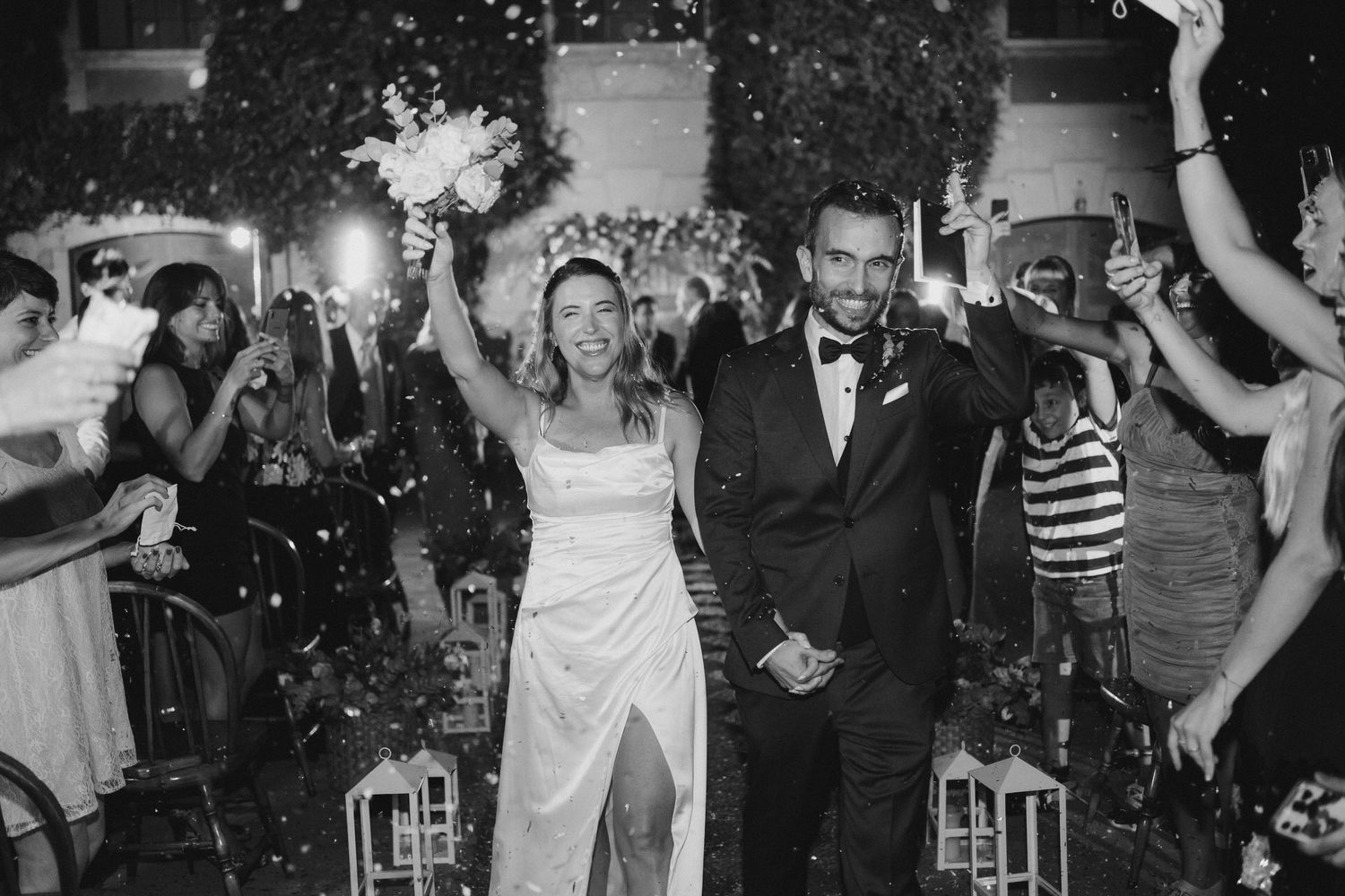 Black and white wedding exit photo of couple celebrating with raised bouquet amid cheering guests at night.