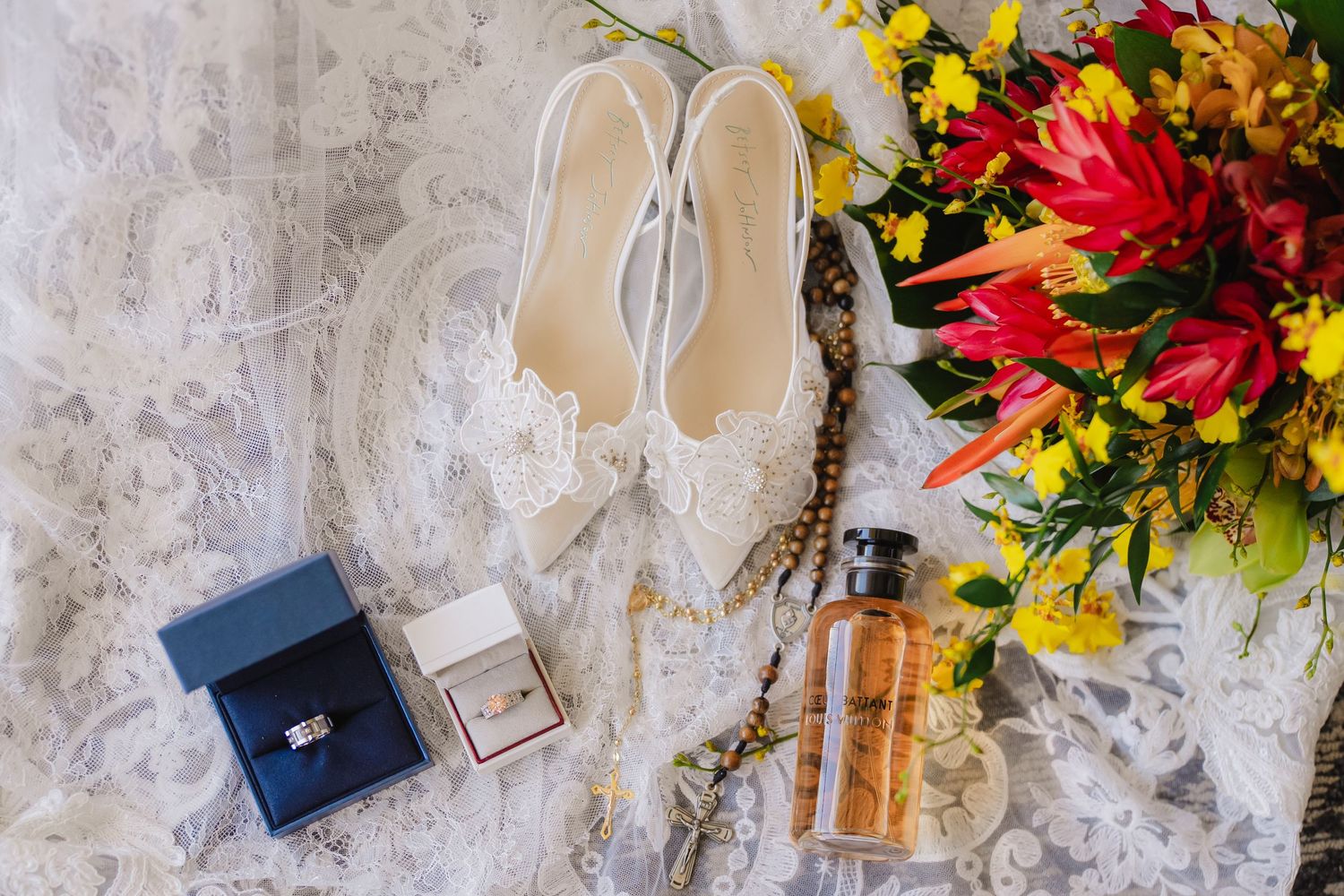 Bridal flat shoes, diamond rings in blue box, perfume bottle, and vibrant tropical bouquet arranged on white lace wedding dress.