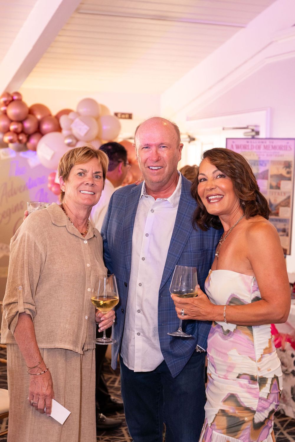 Three guests smile and hold wine glasses at an elegant party with balloon decorations in the background.