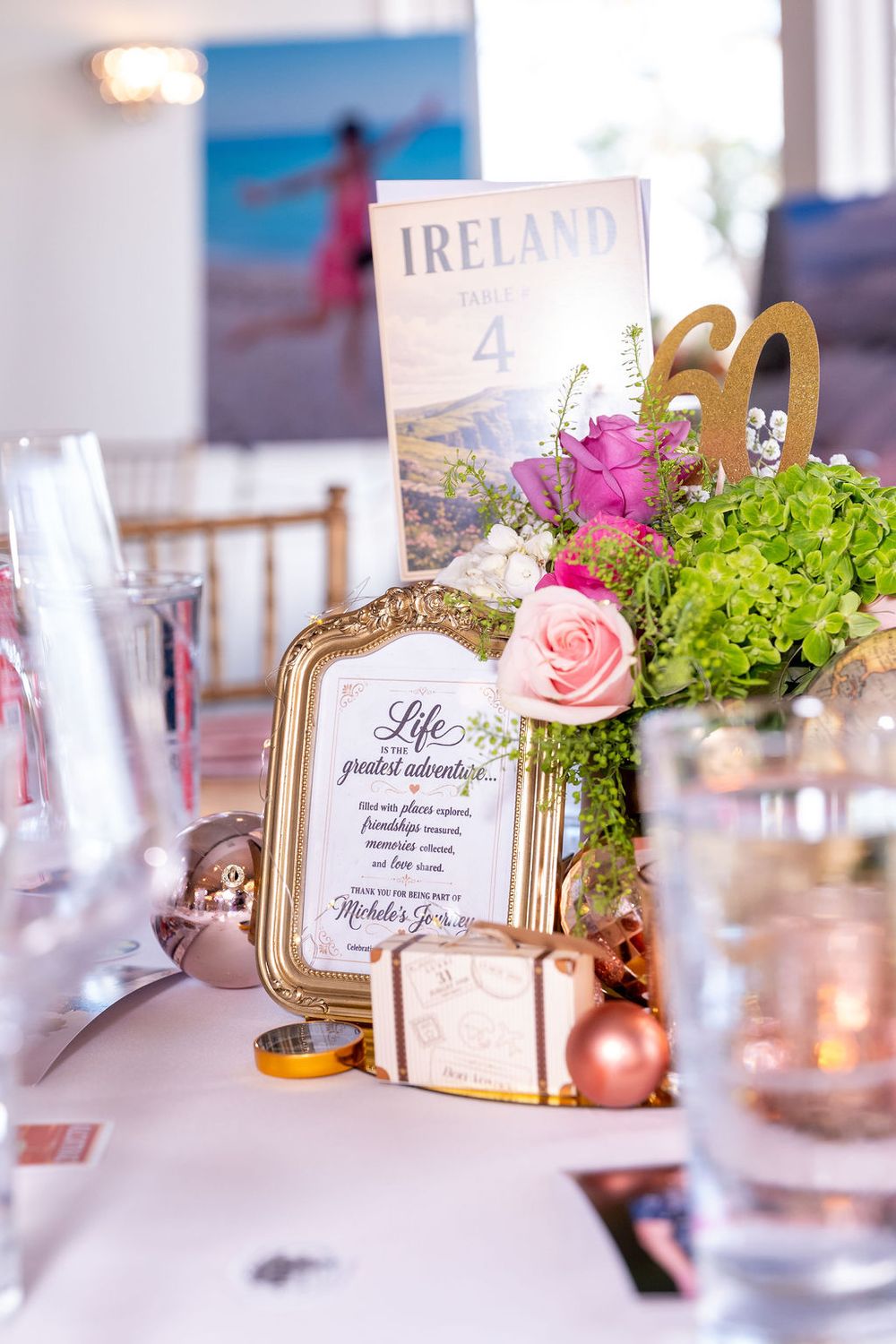 Elegant travel-themed party table centerpiece with Ireland sign, pink roses, green hydrangeas, framed quote, and gold 60th birthday decor.