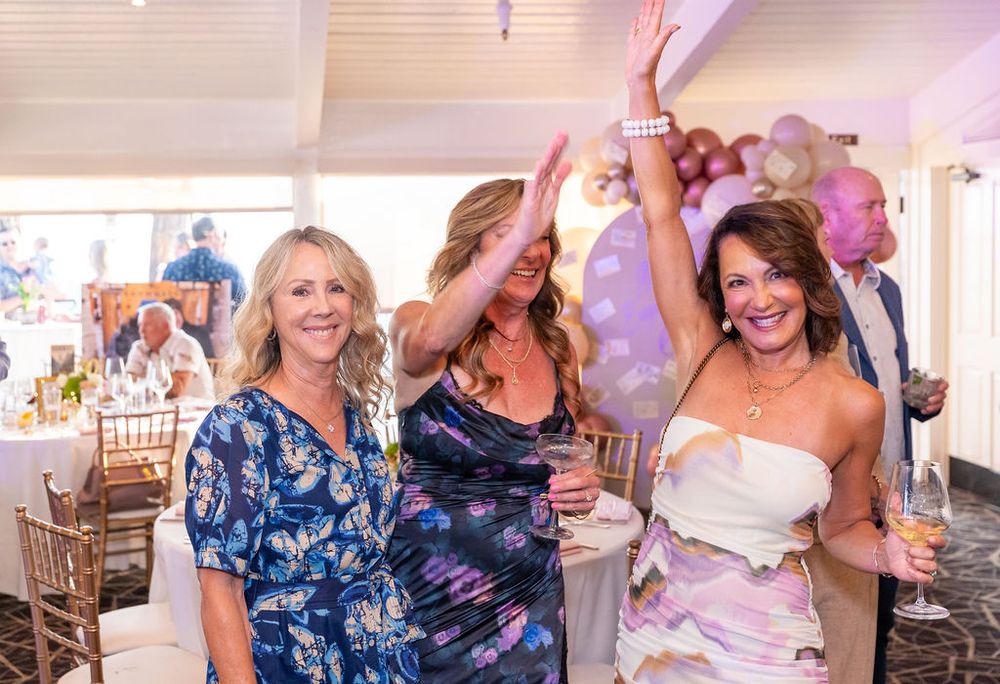 Three women celebrate joyfully at an elegant indoor party with balloons and decorations in the background.