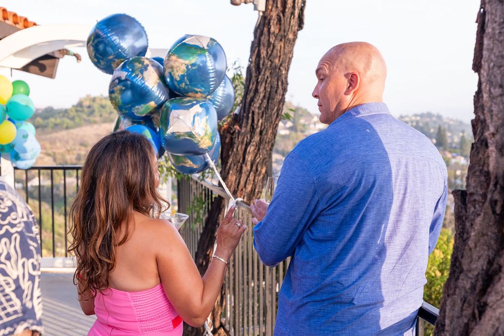 A bald man in a blue shirt and a woman in pink hold globe balloons at an outdoor party with scenic views.
