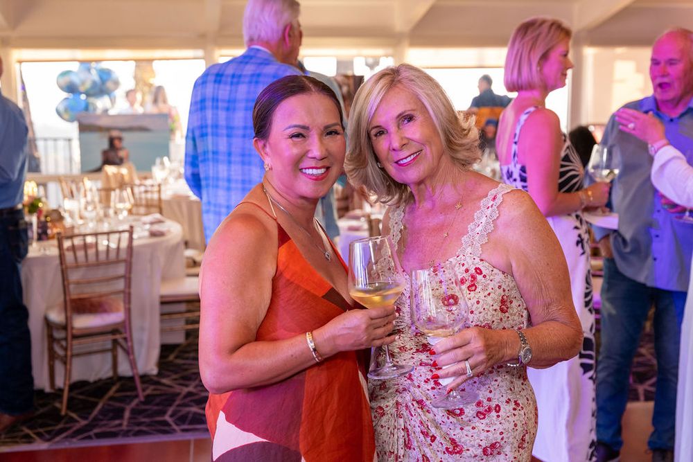Two women smiling and holding wine glasses at an elegant indoor social event or party.