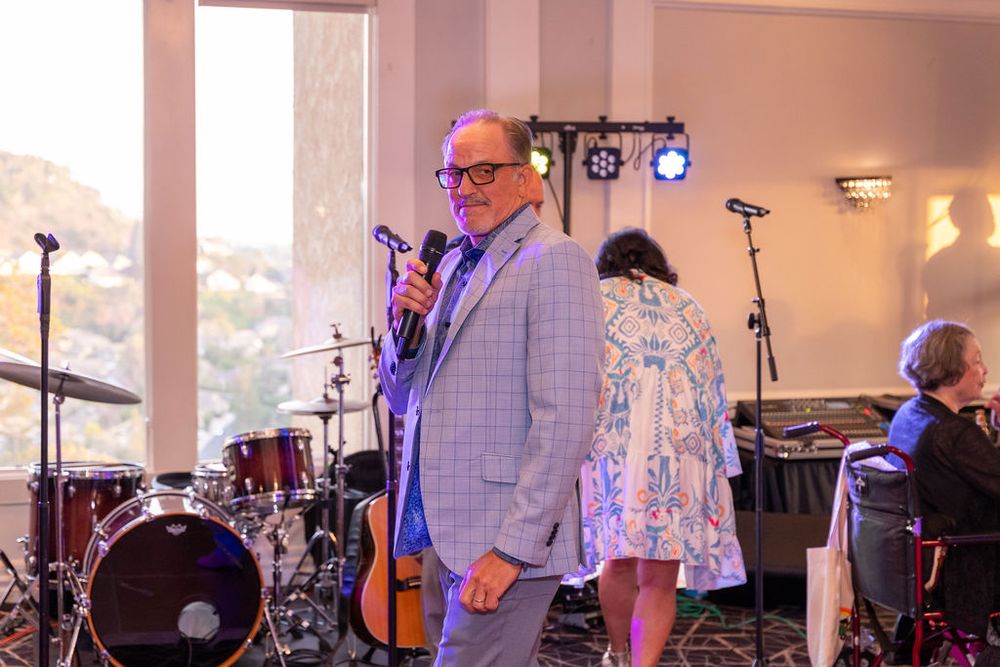 Man in gray blazer holds microphone at indoor event with band setup and guests in background.