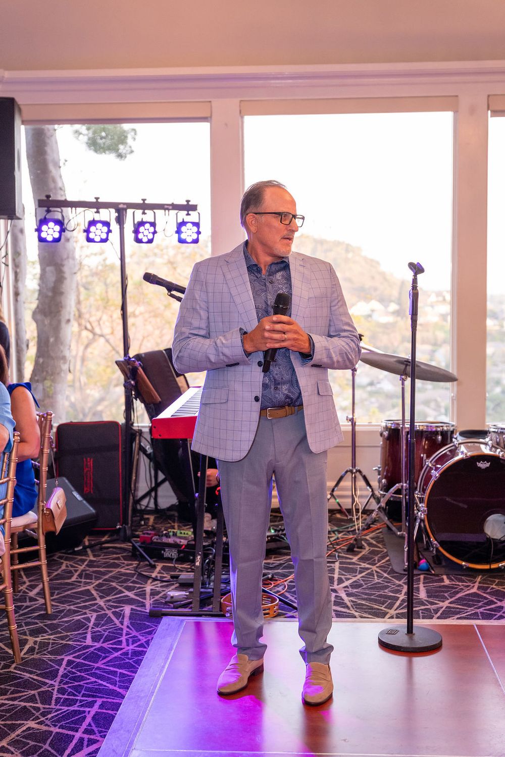 Man in gray suit holding microphone speaks at elegant indoor event with band equipment and large windows behind him.