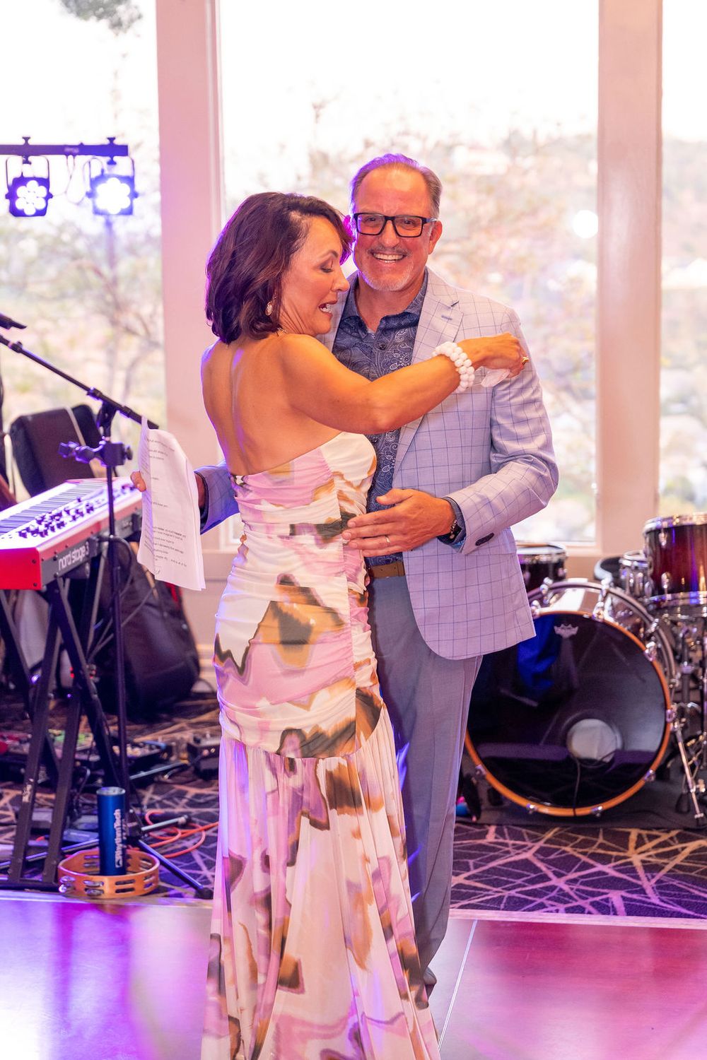 Couple dancing together at an elegant event, woman in floral gown, man in light gray suit, with band instruments in background.