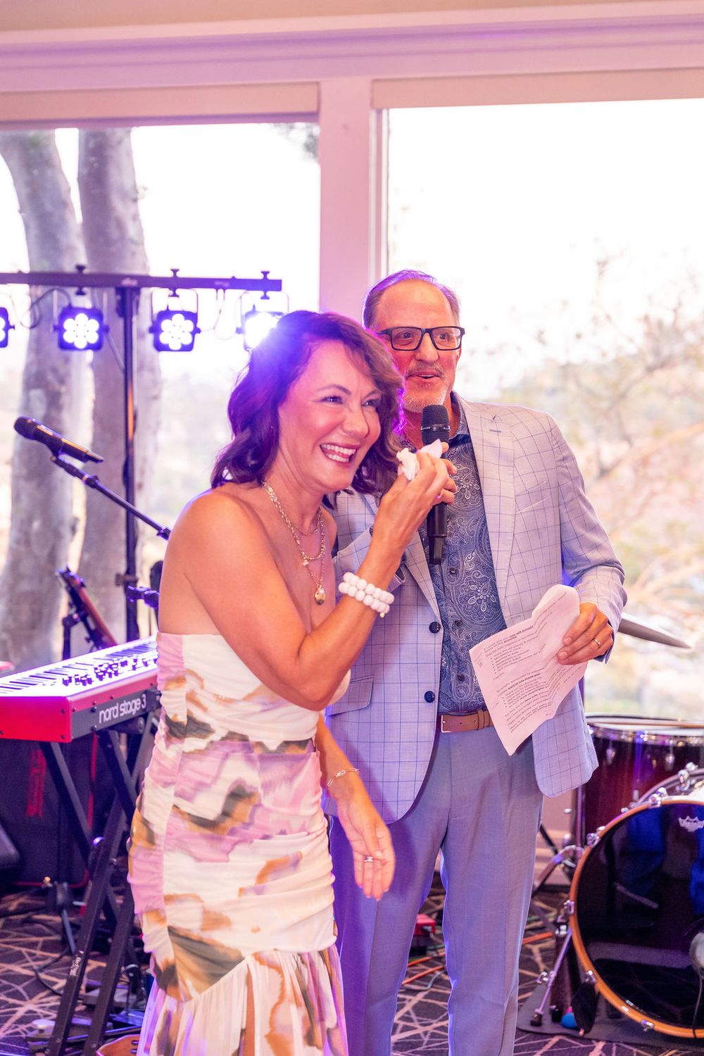 A smiling woman speaks into a microphone beside a man in a suit at an elegant event with band equipment behind them.