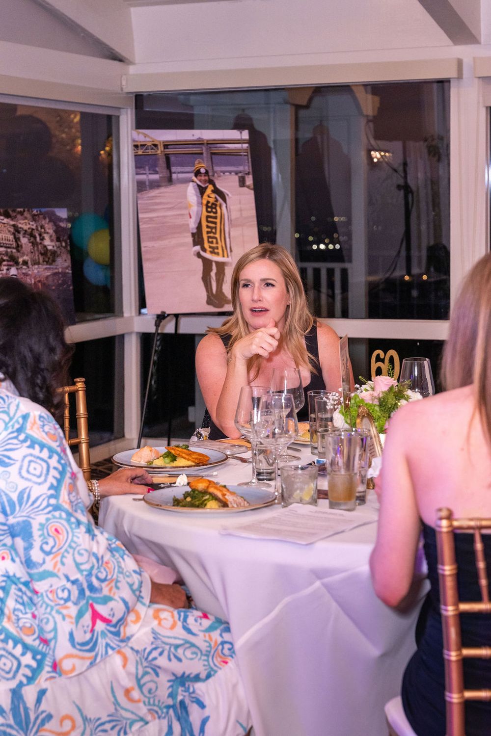 Woman in black dress sits at elegant dinner table during a 60th birthday celebration with a presentation screen in background.