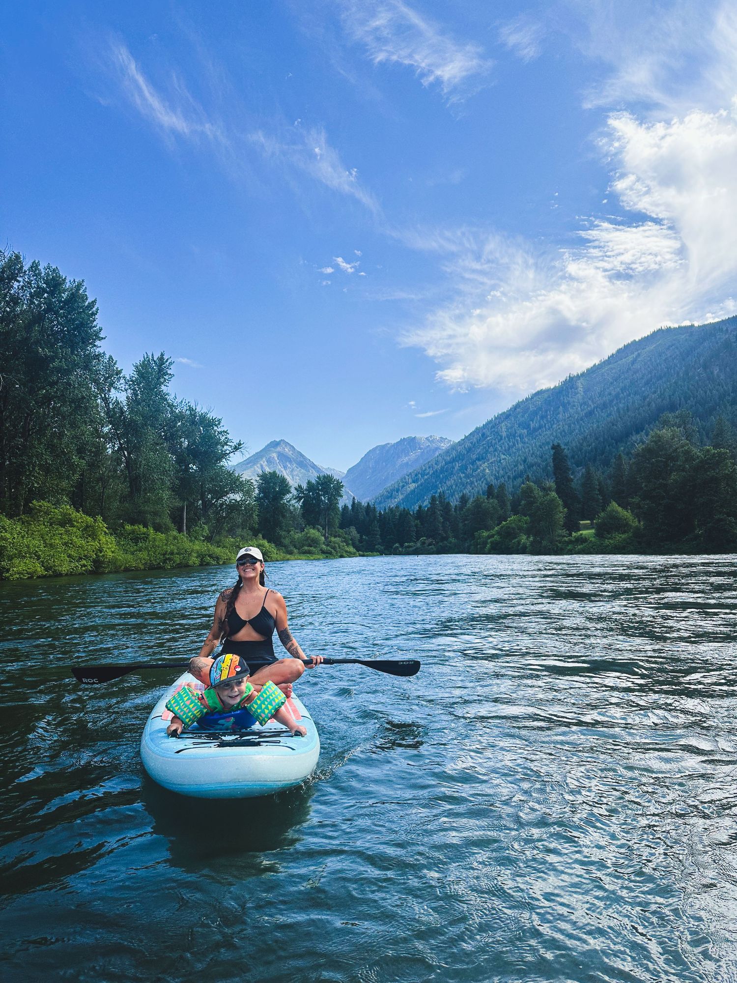 Woman and child paddleboarding on scenic mountain river under blue sky.