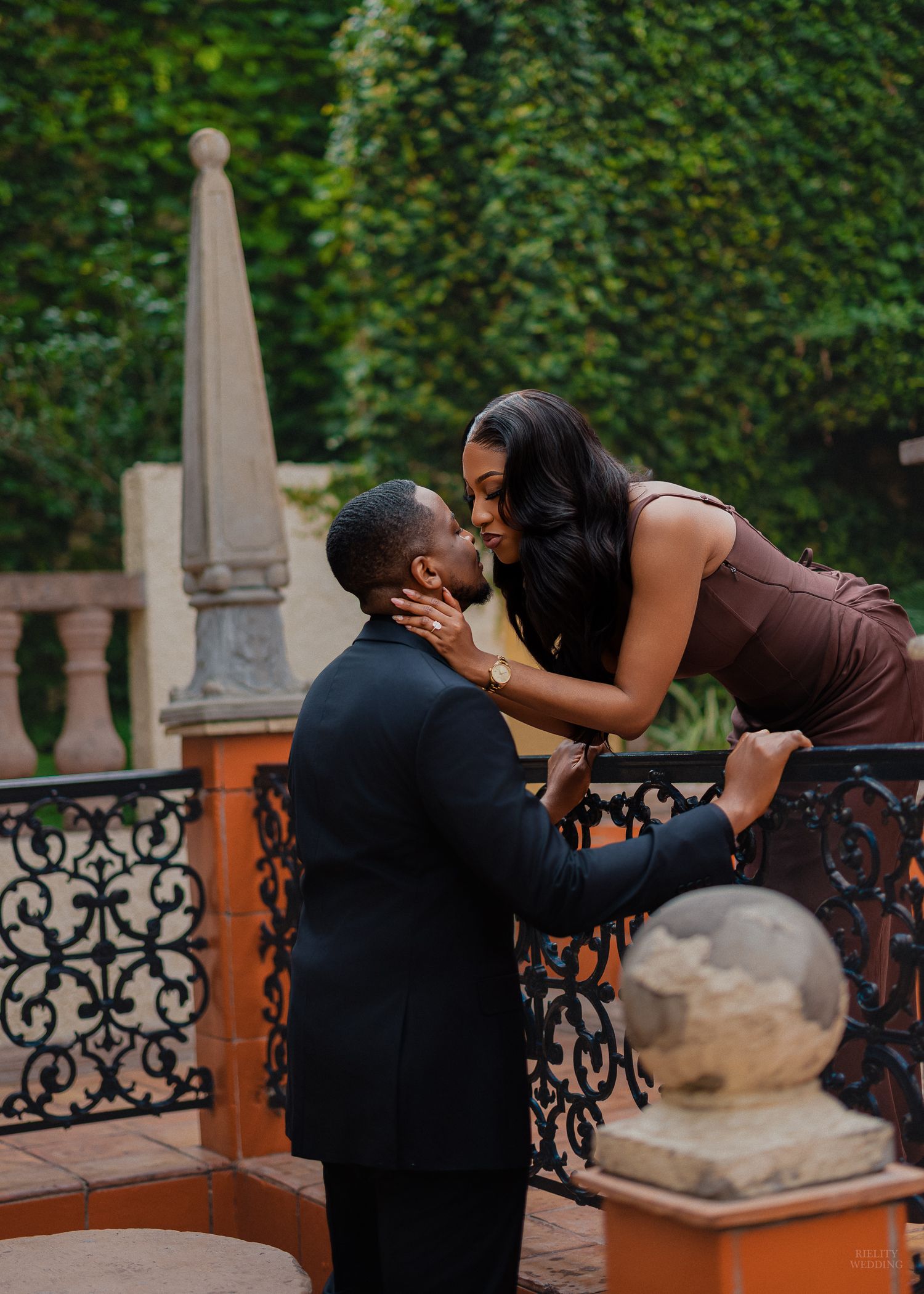 A couple shares a romantic embrace as the man lifts the woman in a sunlit courtyard with stone columns and ivy.