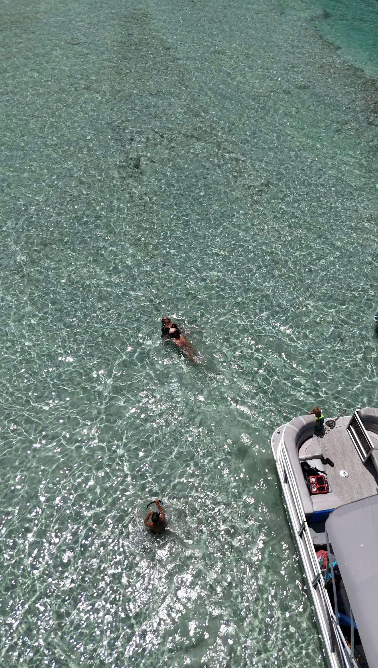 Aerial view of two people snorkeling in crystal-clear shallow turquoise water near a white boat.
