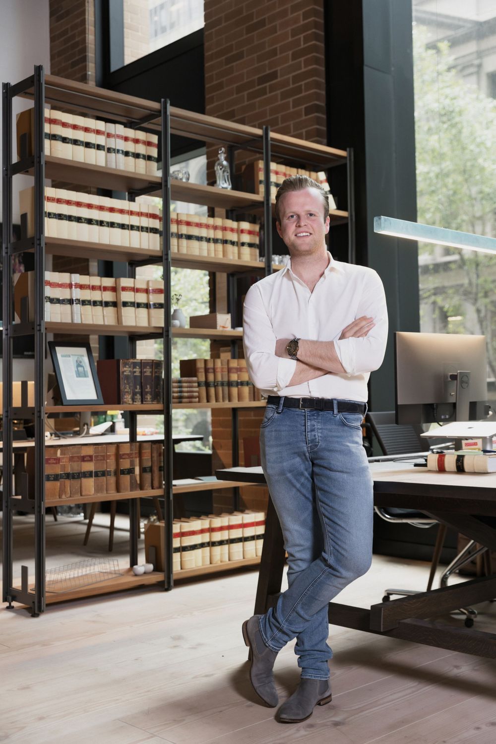 Young professional man in white shirt and jeans leans against bookshelf in stylish office library.