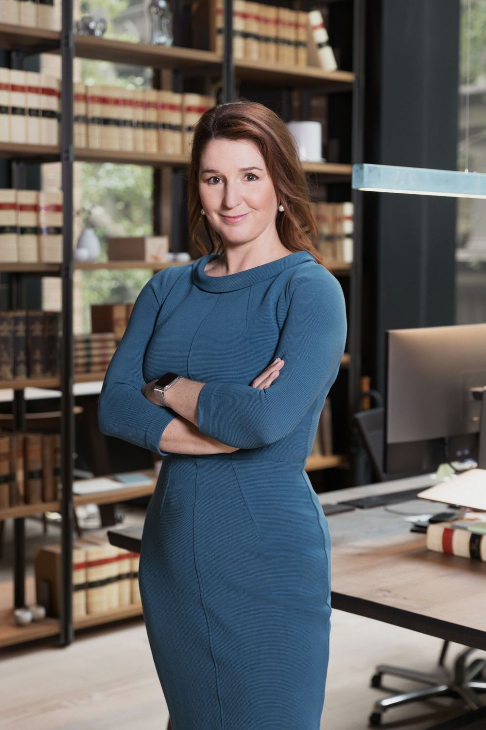Confident businesswoman in teal dress stands with arms crossed in a professional law office library.