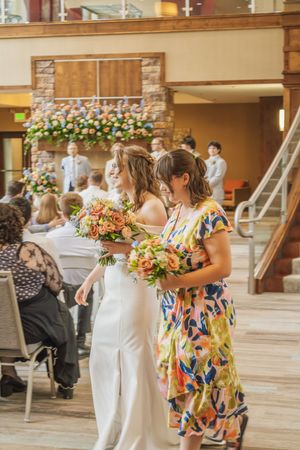 Bride in white gown walks down aisle with bridesmaid holding colorful bouquets at indoor wedding ceremony.