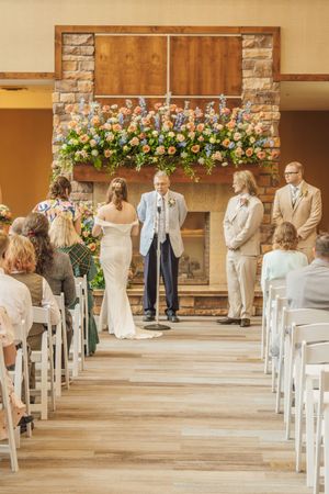 Wide view of indoor wedding ceremony with guests seated in white chairs facing couple at floral altar.