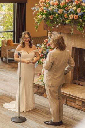 Bride in off-shoulder gown smiles at groom during indoor wedding vows beside large floral arrangement.