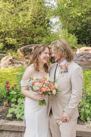 Bride and groom smiling at each other in a lush garden, bride holding a vibrant peach floral bouquet.
