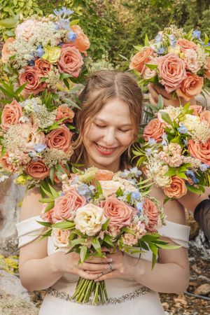 Smiling bride surrounded by bridesmaids holding matching peach and blue rose wedding bouquets outdoors.