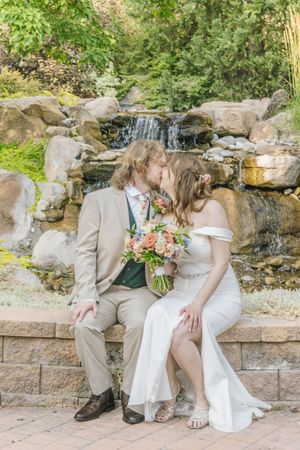 Newlyweds sharing a romantic kiss while seated near a stone waterfall feature in a garden setting.