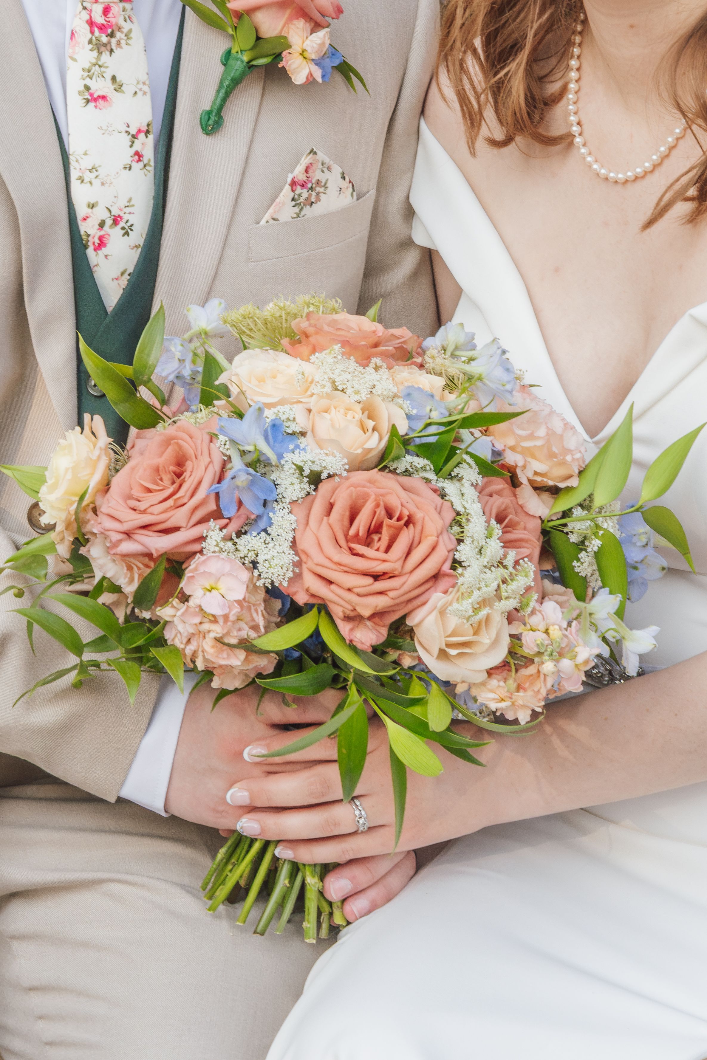 Close-up of bride and groom holding peach rose bouquet with blue flowers, groom in tan suit.