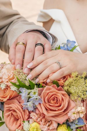 Newlyweds display wedding rings resting hands on peach rose and blue flower bridal bouquet.