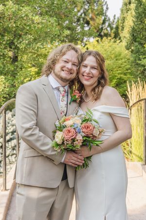 Smiling bride and groom posing together outdoors holding a colorful bouquet of peach and pink roses.