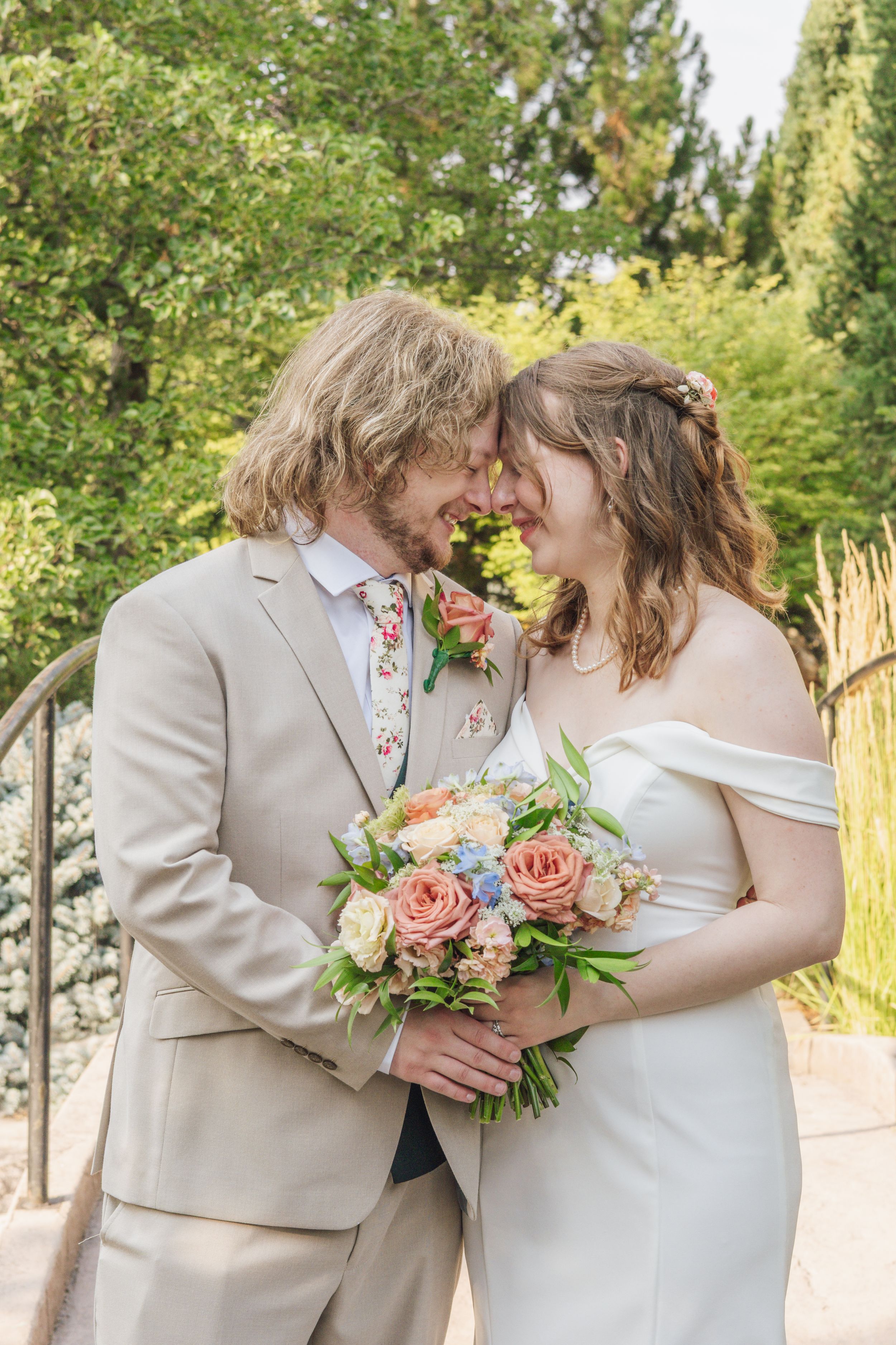 Bride and groom touching foreheads romantically on a garden bridge while holding a pastel floral bouquet.