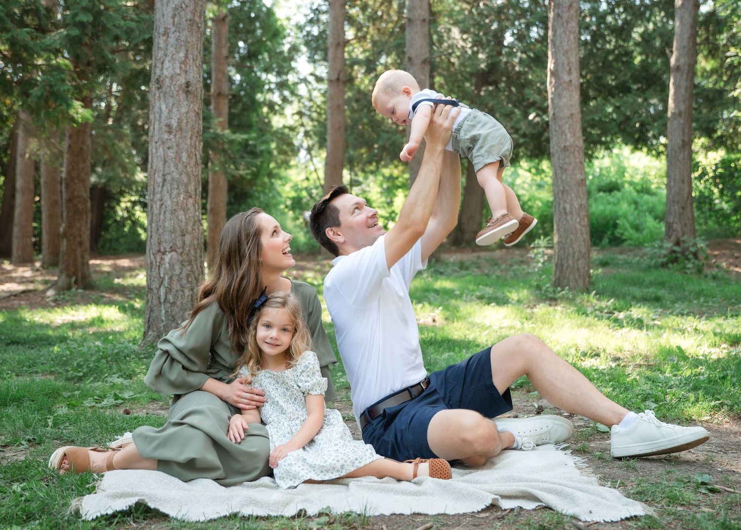 Family enjoying outdoor picnic in forest, parents playing with baby and toddler on blanket among tall trees.
