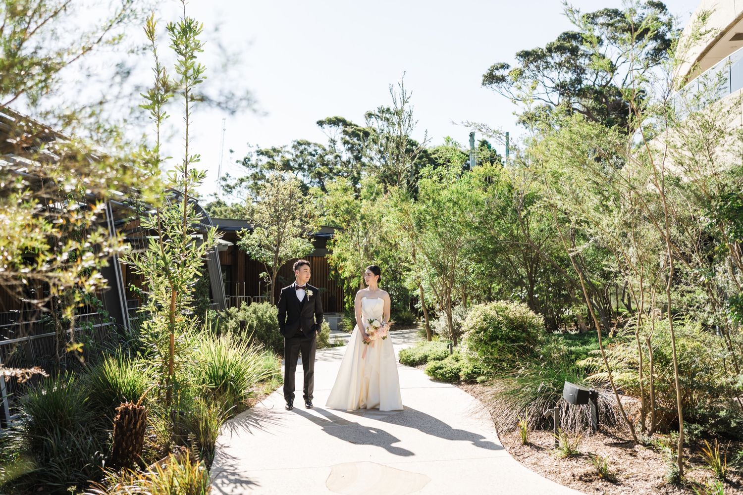 Bride and groom stand on a garden path surrounded by lush greenery and trees on their wedding day.