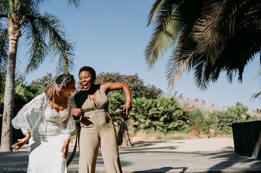 Two women laughing and dancing together outdoors near palm trees on a sunny day at a tropical wedding celebration.