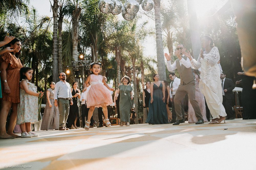 A young girl in a pink dress dances joyfully at an outdoor wedding reception surrounded by guests and palm trees.