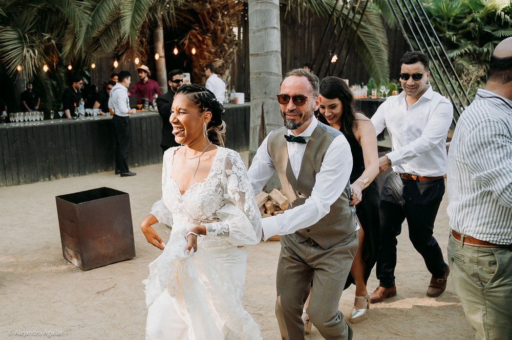 Joyful bride in lace gown and groom in vest and bow tie walking together at an outdoor beach wedding reception.