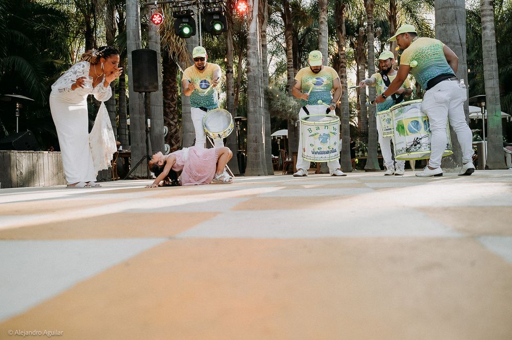 Capoeira performers and drummers in colorful outfits perform outdoors on a decorated stage surrounded by palm trees.