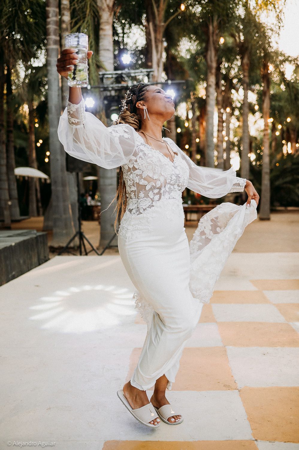 A joyful bride in a white lace jumpsuit dances on a checkered floor at an outdoor wedding reception.