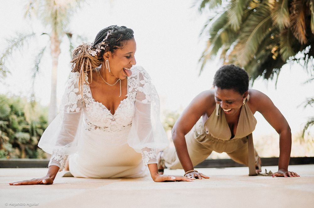 Bride in white lace dress laughing with woman doing push-ups on outdoor patio near palm trees.