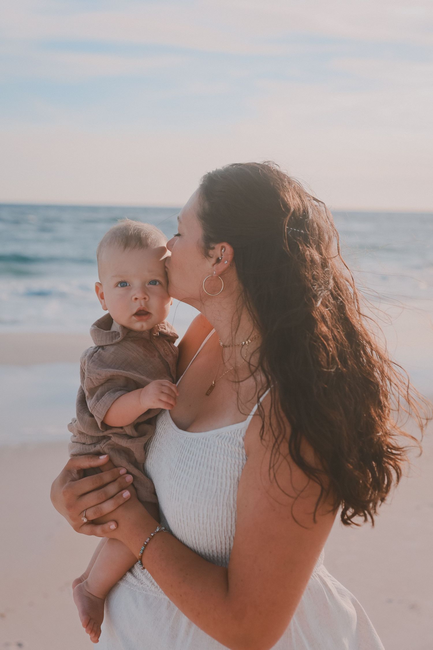 A mother tenderly kisses her baby on the cheek while standing on a beach, both dressed casually near ocean waves.