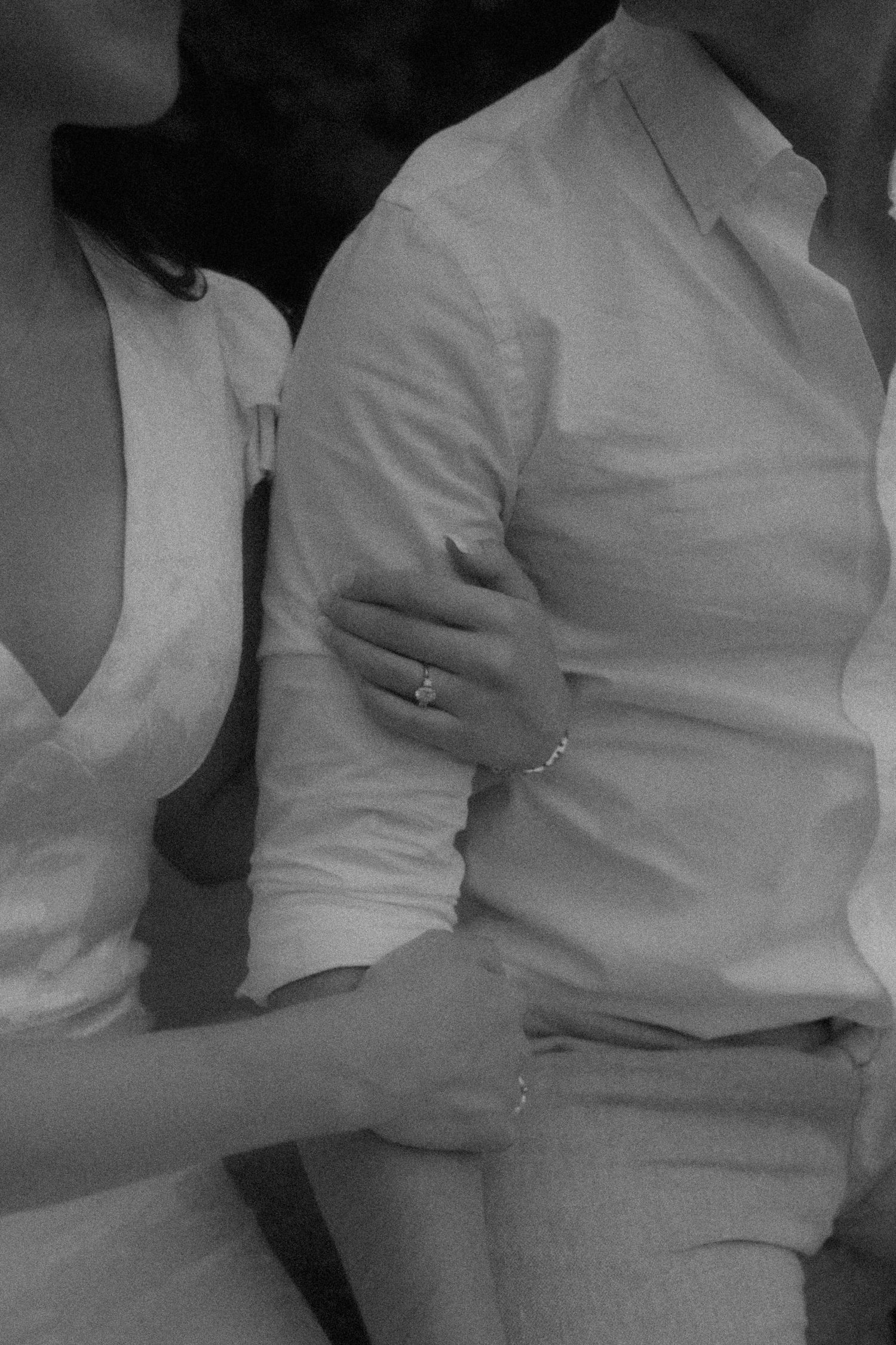 Black and white photo of a couple embracing, woman wearing an engagement ring and bracelet, both dressed in white. Whistling Gardens Ontario