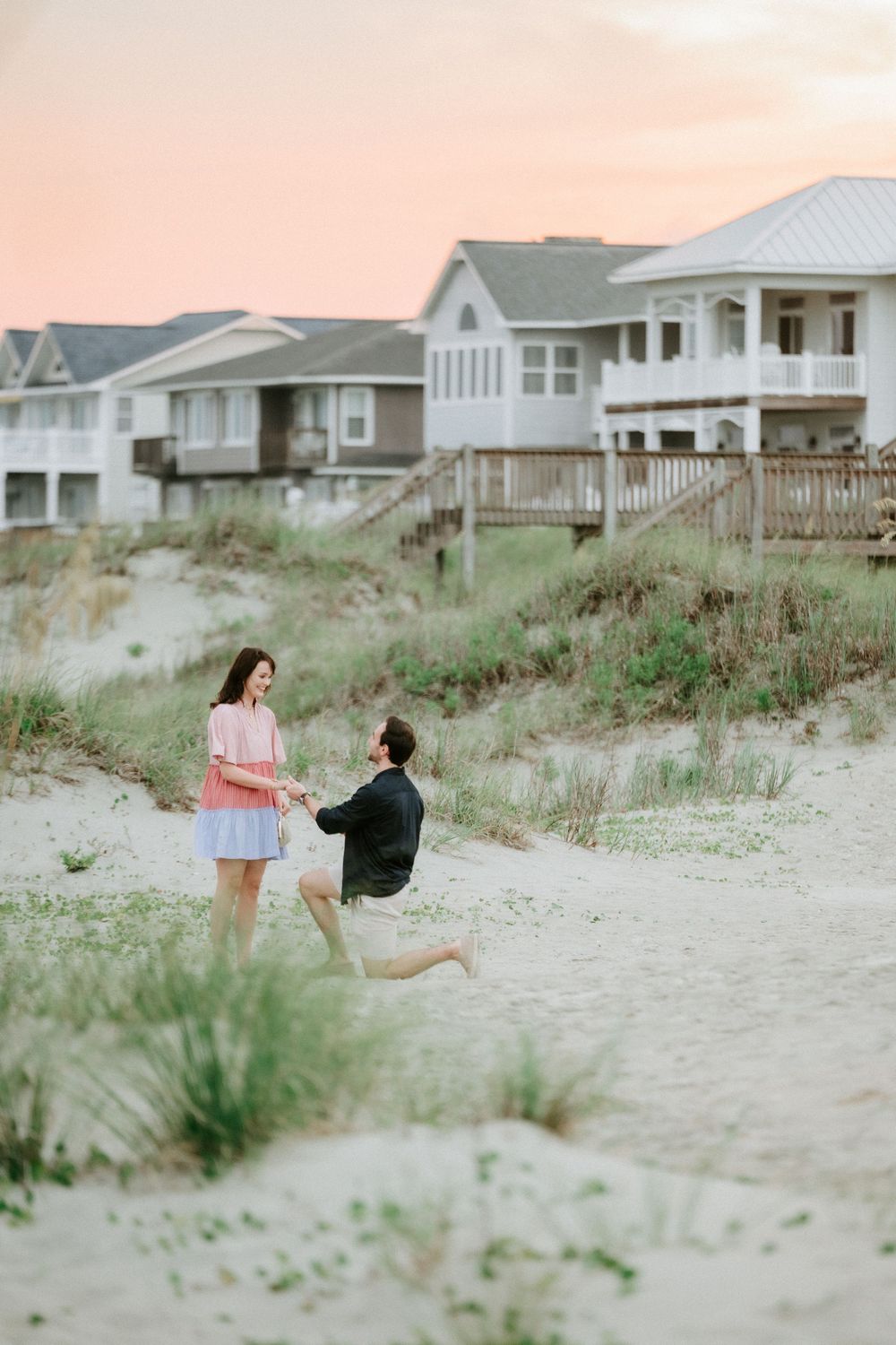 Man proposes on one knee to woman on sandy beach at sunset with coastal houses in background.