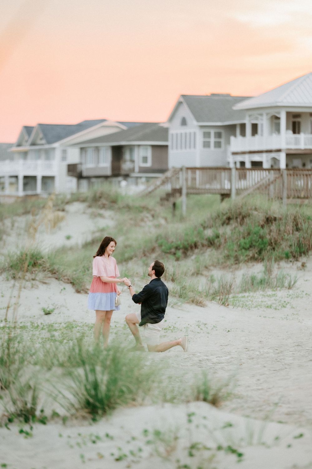 Man proposes on sandy beach at sunset with coastal houses in background, woman in pink dress reacts with joy.