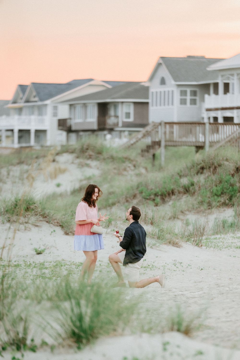 Man proposes on sandy beach at sunset near coastal houses, kneeling before woman in pink dress.