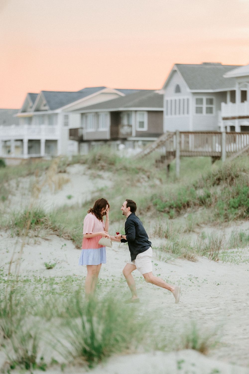 Couple sharing a joyful moment on sandy beach dunes at sunset with coastal houses in the background.