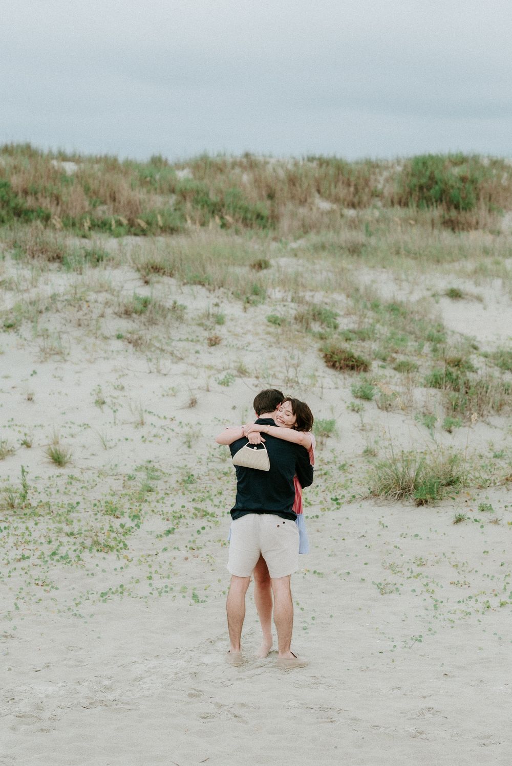Couple embracing on sandy beach dunes with green grass and overcast sky in background.