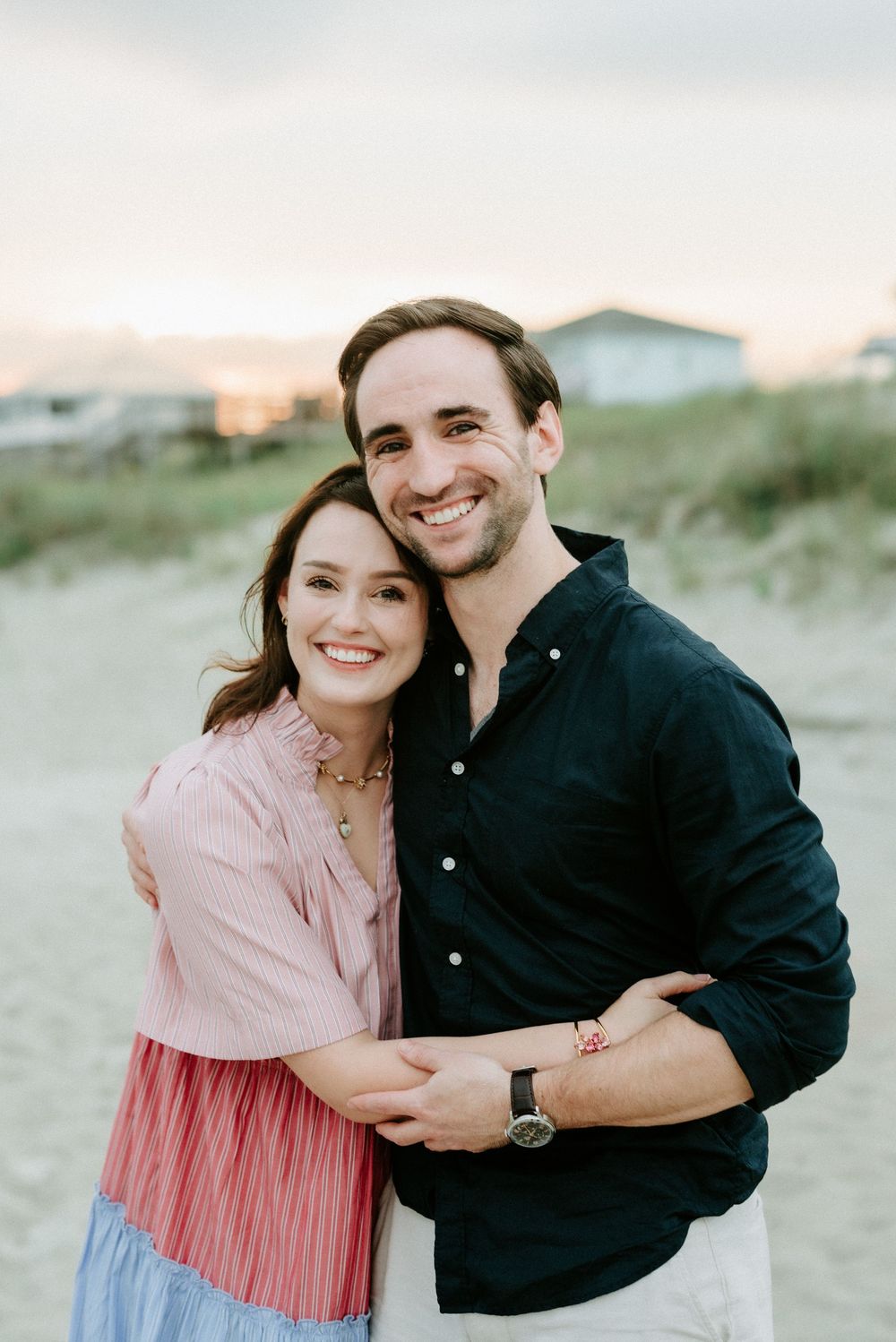 Happy couple embraces on beach at sunset, woman in pink dress and man in navy shirt smiling warmly.