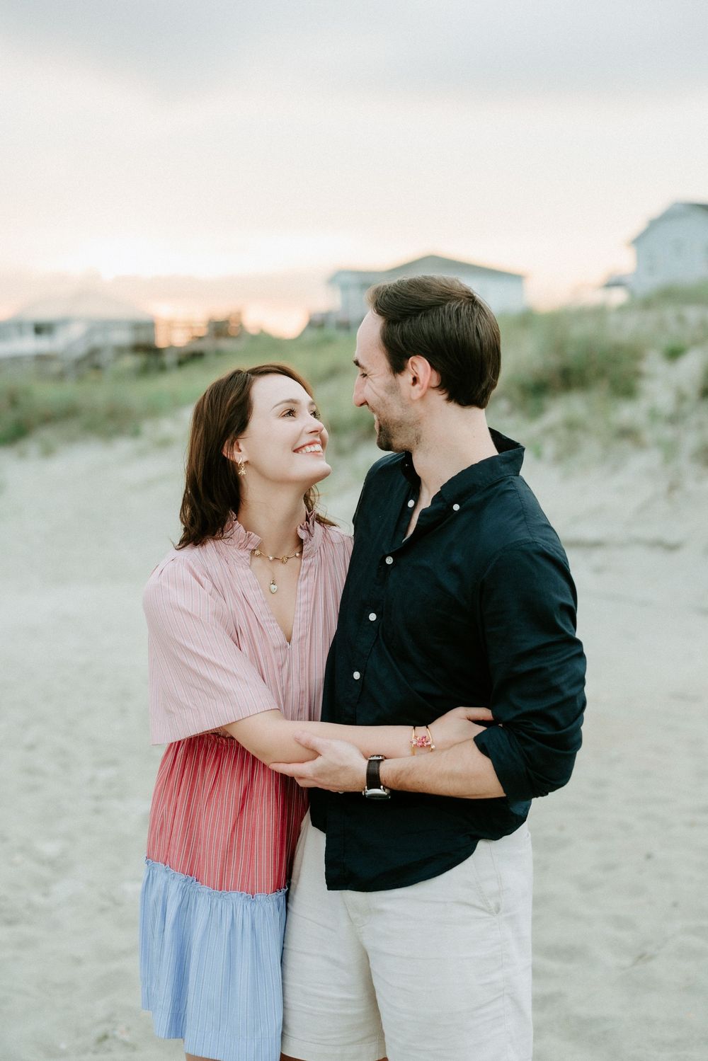 A smiling couple embraces on a sandy beach at sunset, the woman in a pink dress gazing up at the man in a dark shirt.