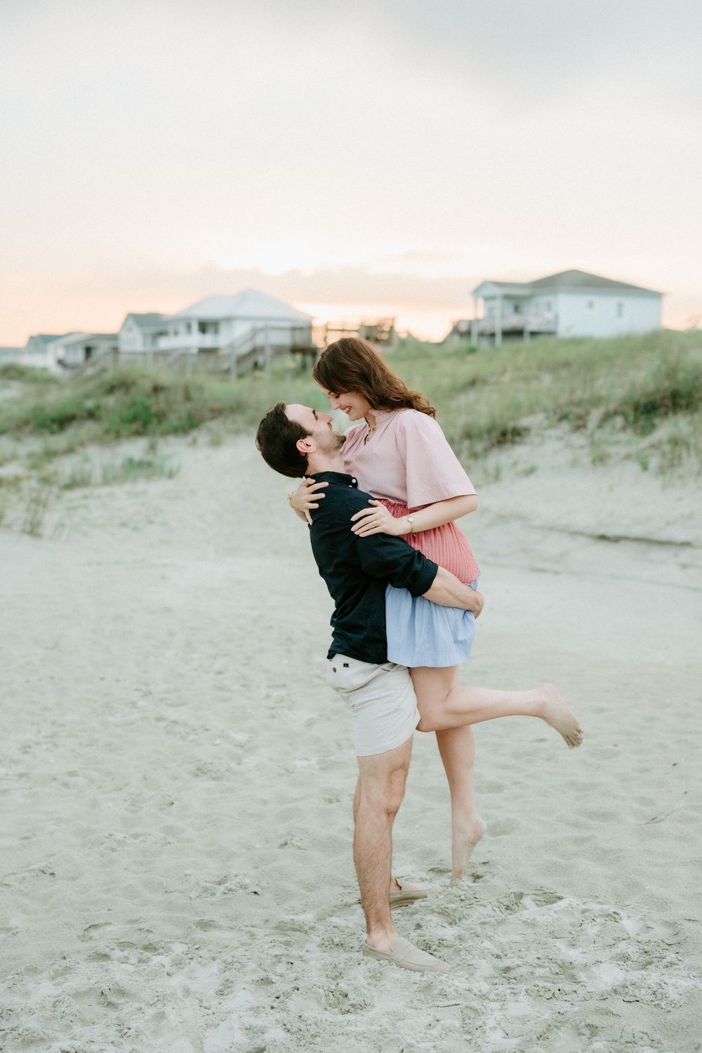 Couple sharing a romantic kiss on sandy beach at sunset, man lifting woman near coastal dunes.