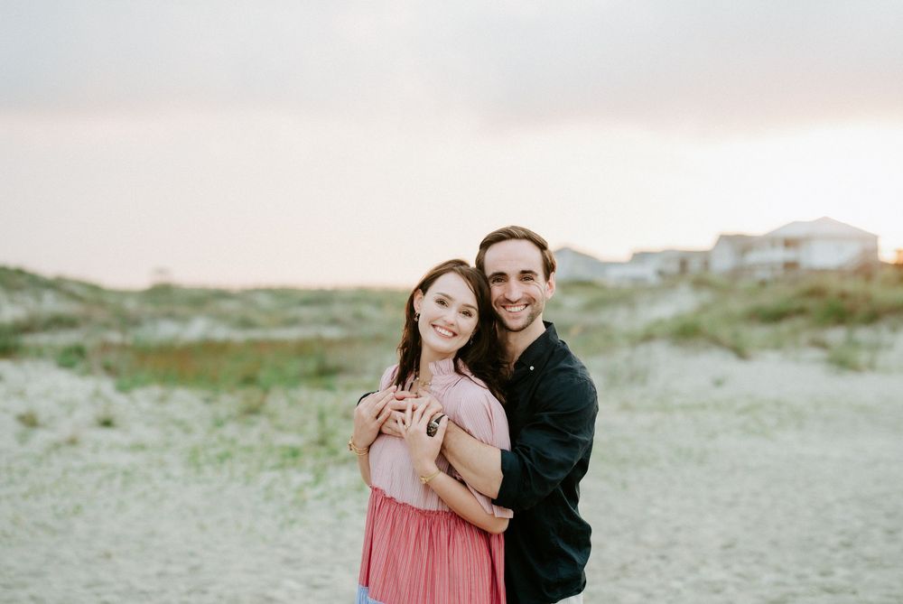 A couple embraces and smiles on a sandy beach at sunset, woman in pink dress, man in dark shirt.