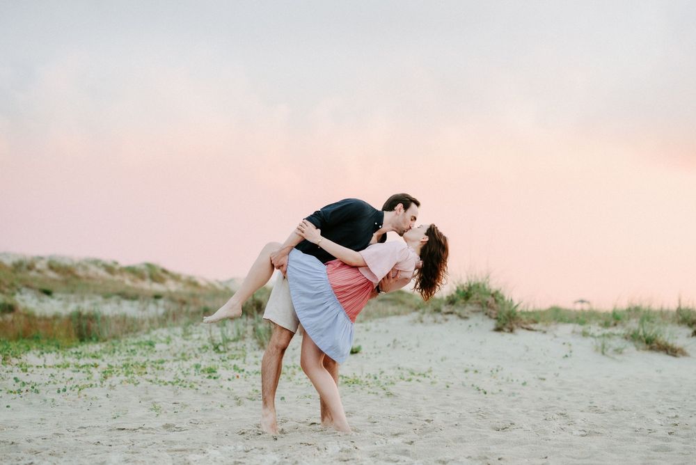 Couple sharing a romantic dip kiss on a sandy beach at sunset with pink sky and dunes in the background.