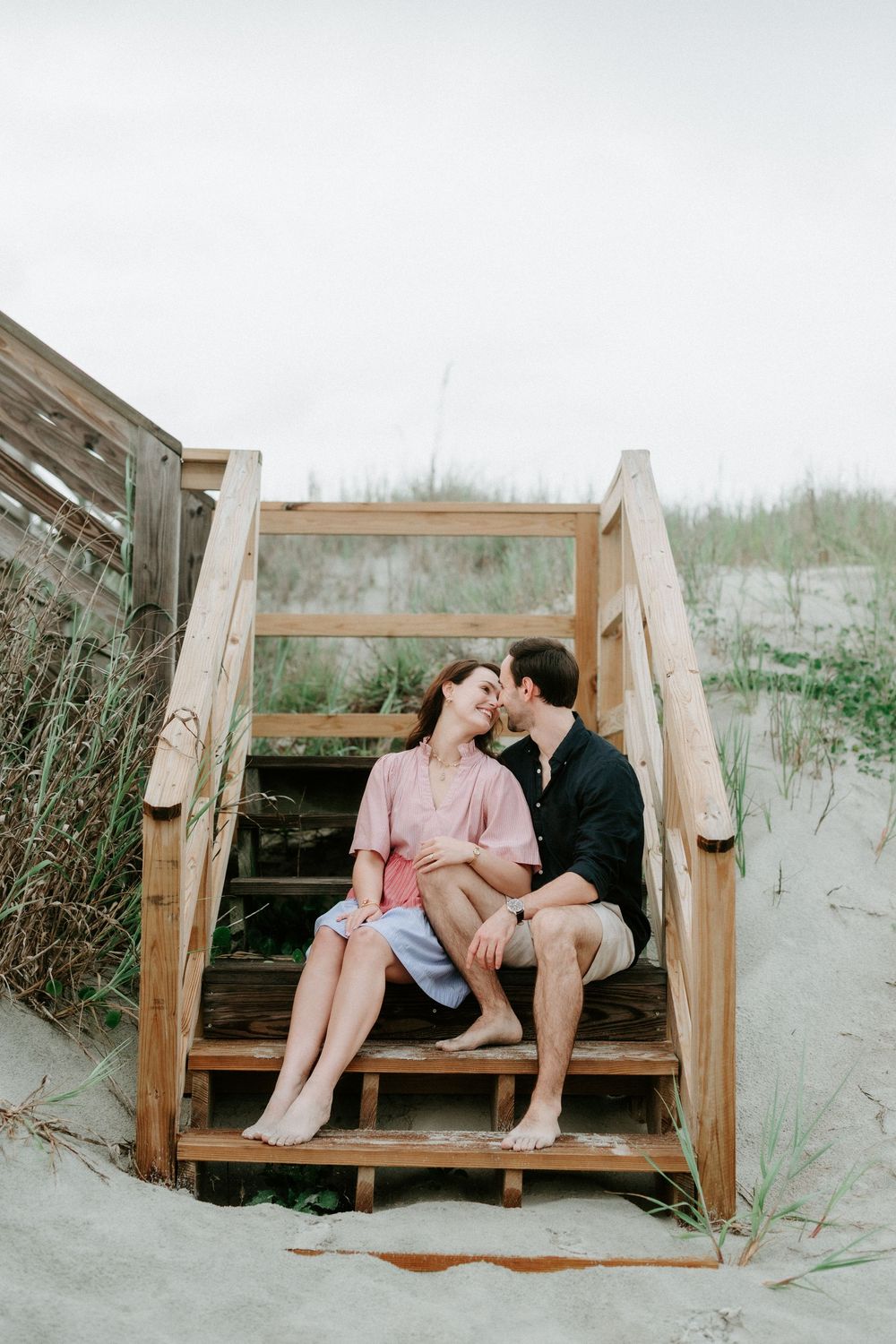 A couple sits closely on wooden beach stairs, sharing a tender kiss surrounded by sand dunes and coastal grass.