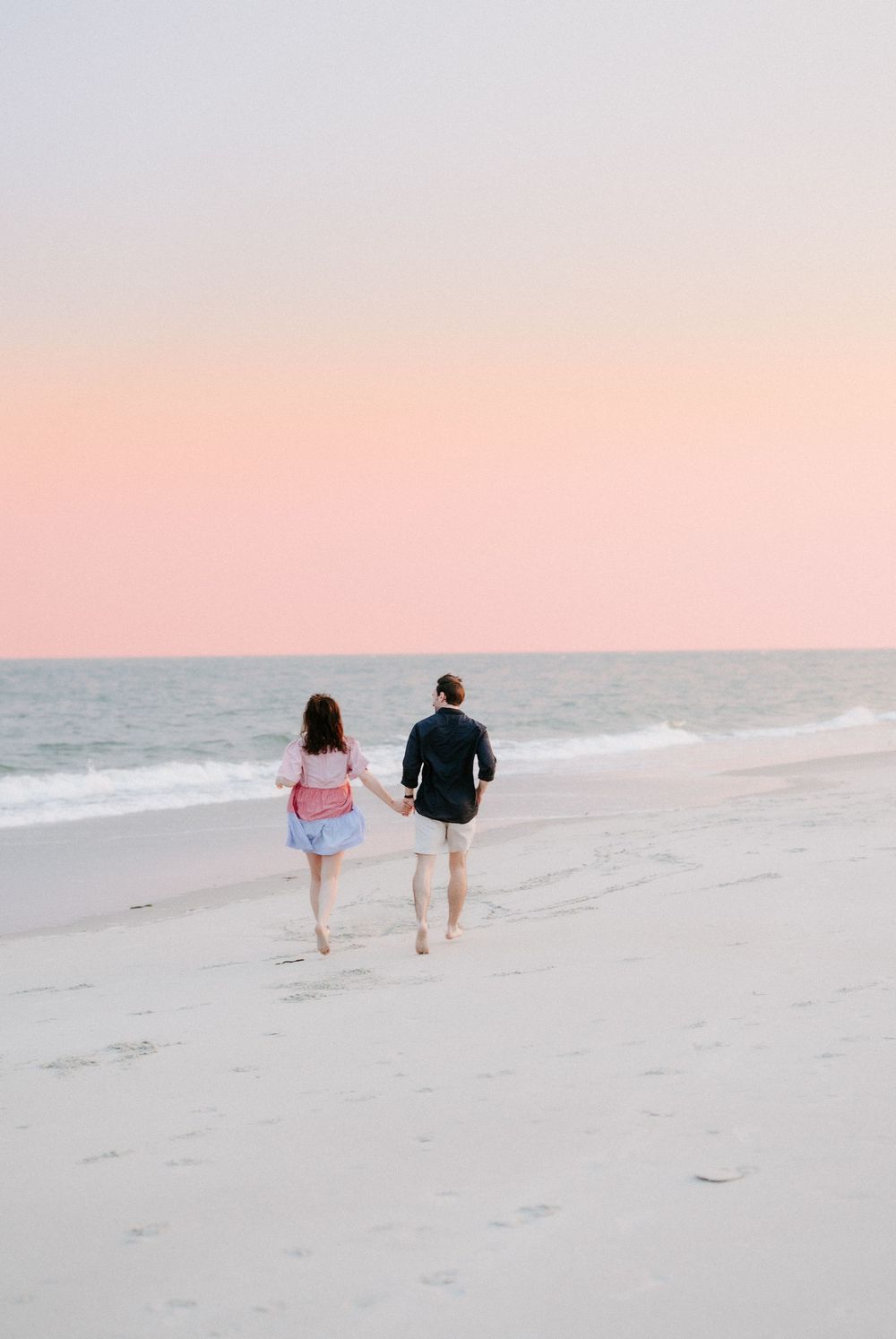 Couple holding hands walking barefoot on a sandy beach at sunset with a pink pastel sky.